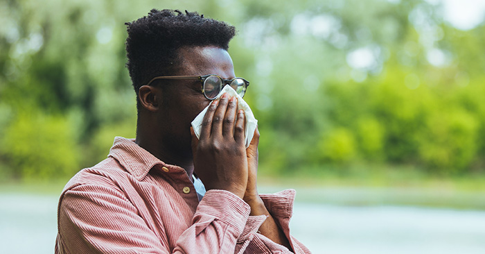 A man blowing his nose into a tissue outside due to outdoor allergens.